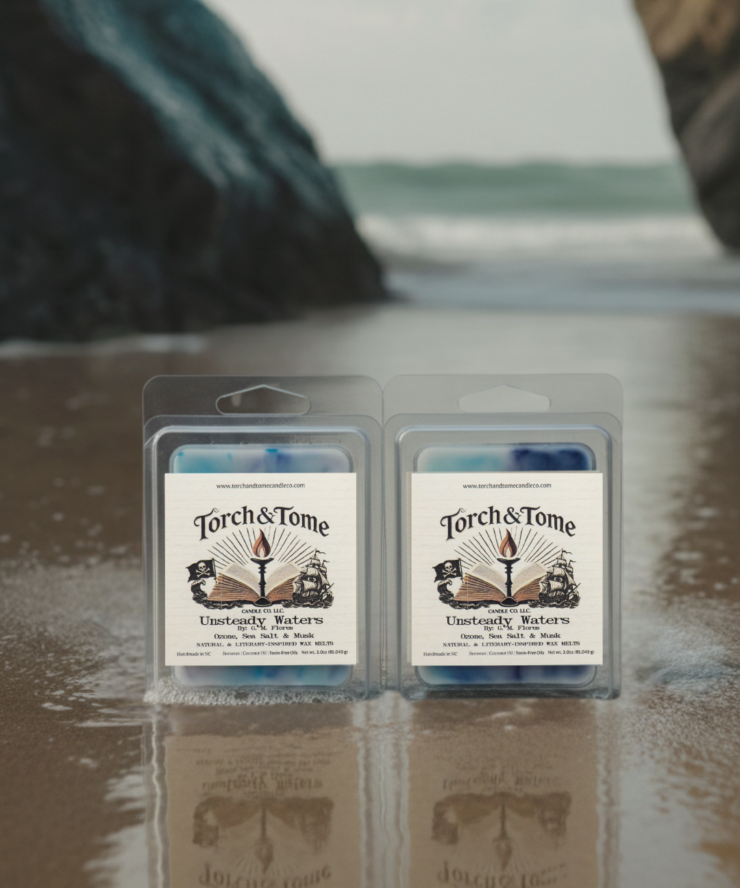Two packages of Torch & Tome clamshells featuring blue, white, and violet marbled wax melts on a reflective surface with a blurred natural washed ashore beach background