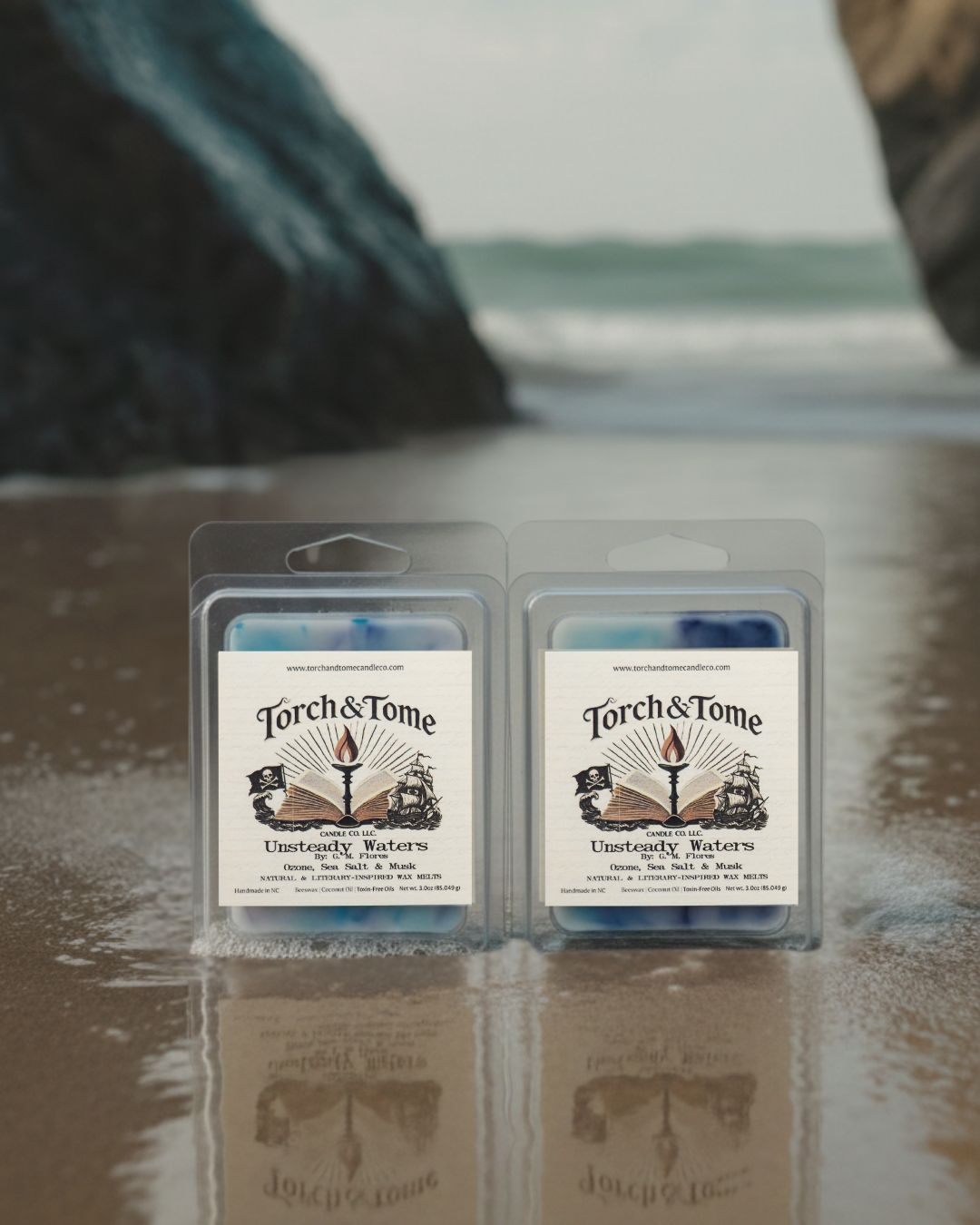 Two packages of Torch & Tome clamshells featuring blue, white, and violet marbled wax melts on a reflective surface with a blurred natural washed ashore beach background