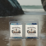 Two packages of Torch & Tome clamshells featuring blue, white, and violet marbled wax melts on a reflective surface with a blurred natural washed ashore beach background