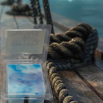 Packaged blue white and violet marbled wax melts on a wooden pirate ship deck surface with a rope and blurred ocean background
