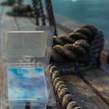 Packaged blue white and violet marbled wax melts on a wooden pirate ship deck surface with a rope and blurred ocean background
