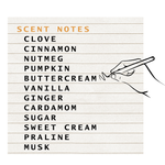 A graphic with a lined paper background reading “SCENT NOTES” in orange, followed by a list of fragrance notes in black: clove, cinnamon, nutmeg, pumpkin, buttercream, vanilla, ginger, cardamom, sugar, sweet cream, praline, and musk. A simple line drawing of a hand holding a pen appears on the right.
