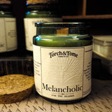 Candle labeled 'Melancholic' by Torch & Tome on a surface with other candles in the background.