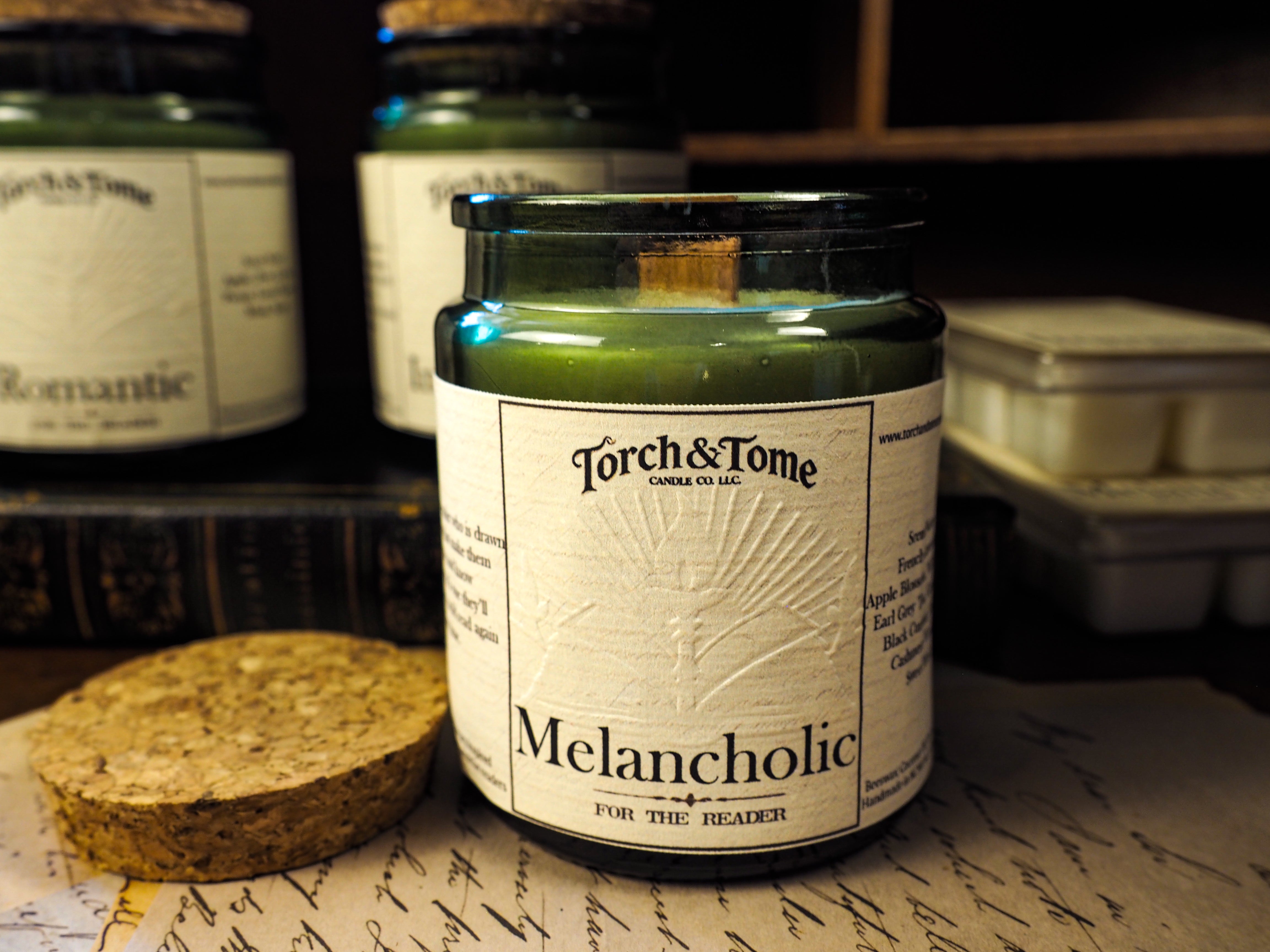 Candle labeled 'Melancholic' by Torch & Tome on a surface with other candles in the background.