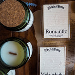 Two boxes of 'Torch & Tome' waxmelts labeled 'Romantic' and 'Melancholic' on a wooden surface with a candle next to them.