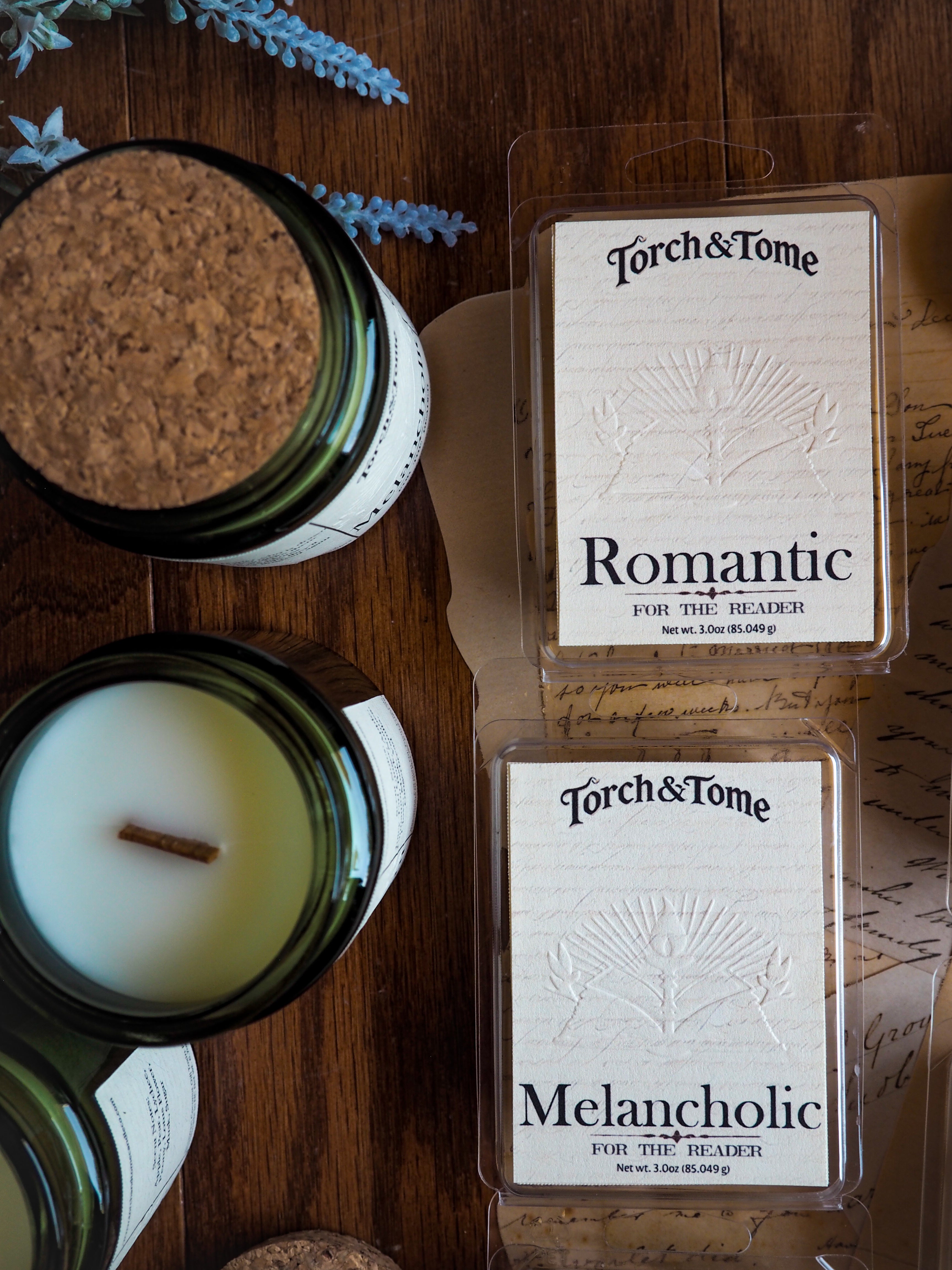 Two boxes of 'Torch & Tome' waxmelts labeled 'Romantic' and 'Melancholic' on a wooden surface with a candle and jar.