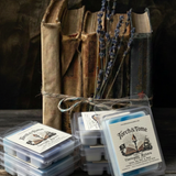 Torch & Tome wax melt clamshells stacked on a rustic wooden table, featuring blue and white wax melts, arranged next to tied vintage books and dried lavender stems.