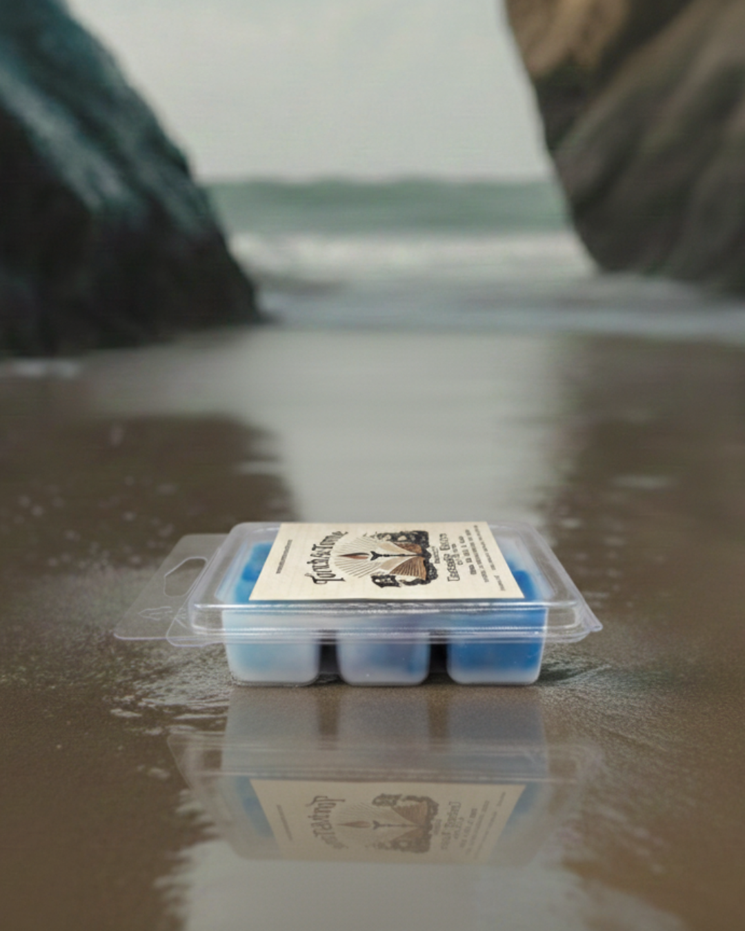 Packaged Torch & Tome clamshell featuring blue white and violet marbled wax melts with a label on a reflective beach surface