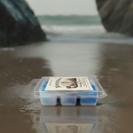 Packaged Torch & Tome clamshell featuring blue white and violet marbled wax melts with a label on a reflective beach surface