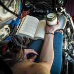 A person sitting on a motorcycle in a garage reads an open book while holding a lit candle in a glass jar. The scene is shot from a high angle, showing the person's arms, hands, and hair, with the motorcycle's chrome details and engine visible in the background.