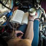 A person sitting on a motorcycle in a garage reads an open book while holding a lit candle in a glass jar. The scene is shot from a high angle, showing the person's arms, hands, and hair, with the motorcycle's chrome details and engine visible in the background.