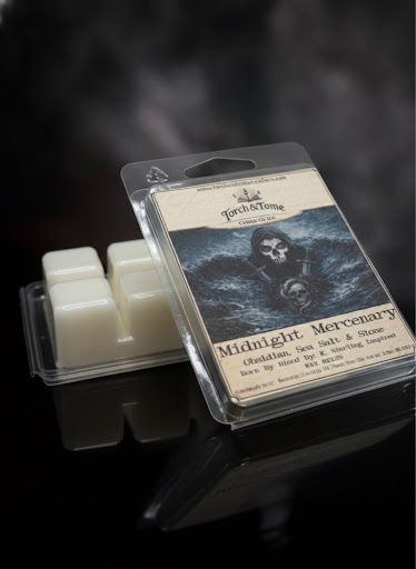 Midnight Mercenary packaged wax melts with a label featuring a skull design and two french swords on the label. Wax melt is propped against an upside down waxmelt clamshell showing the ivory beewax on a dark smoky background