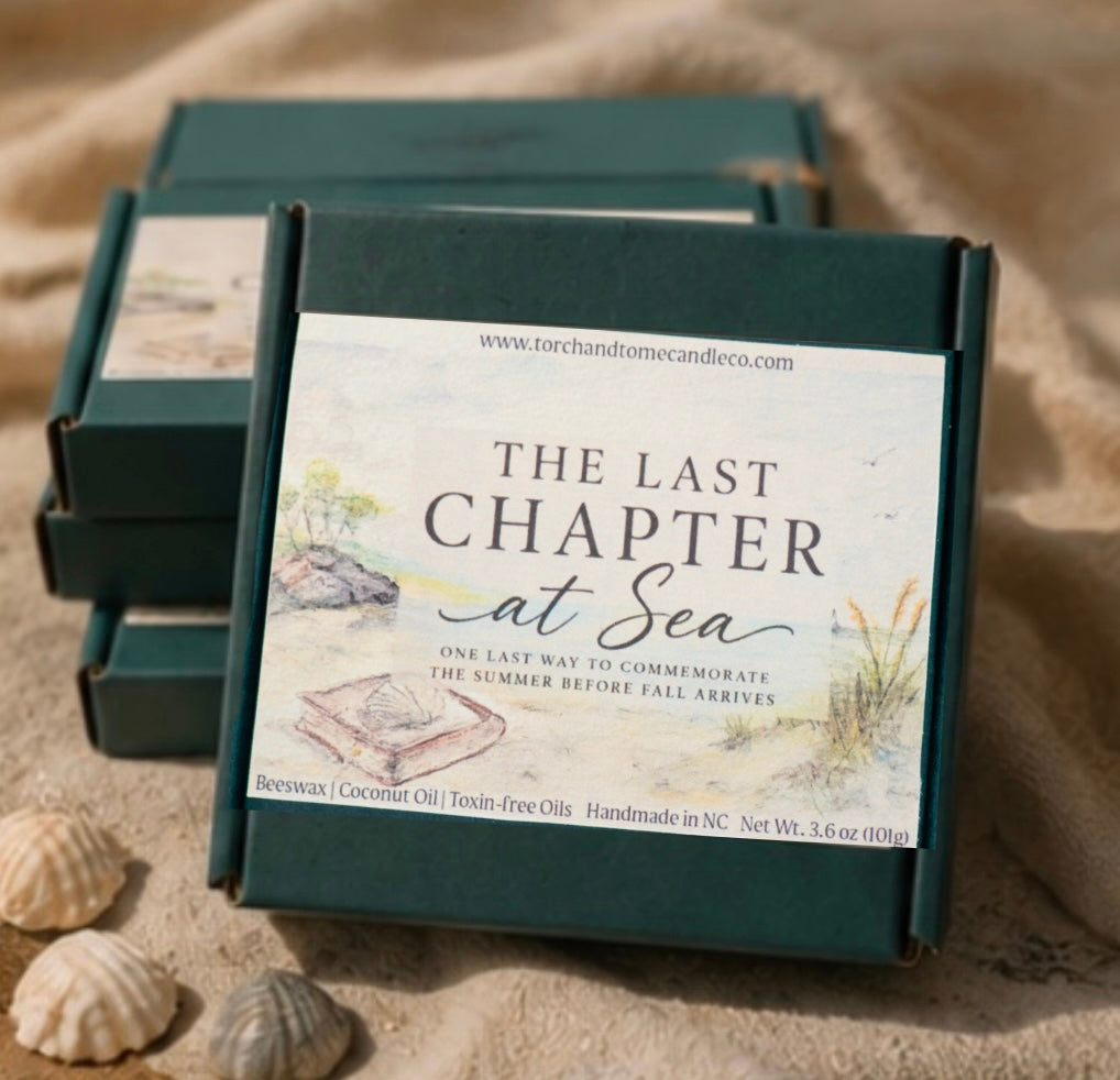 A dark green rectangular wax melt box with a large cream label reading 'THE LAST CHAPTER at Sea' is placed on a sandy surface with shells nearby. Three matching boxes are stacked behind it in a beach-like setting.