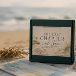 A square dark green gift box labeled 'THE LAST CHAPTER at Sea' stands on a beach-themed surface with shells, dried grass, and a blurred ocean background.