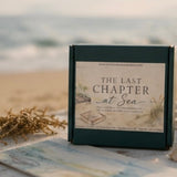 A square dark green gift box labeled 'THE LAST CHAPTER at Sea' stands on a beach-themed surface with shells, dried grass, and a blurred ocean background.