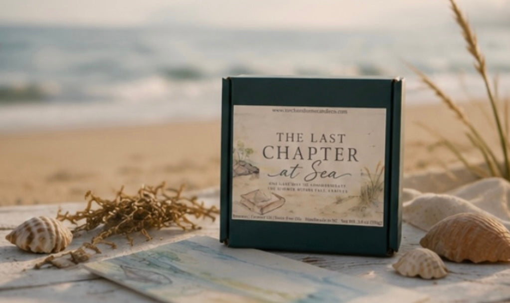 A square dark green gift box labeled 'THE LAST CHAPTER at Sea' stands on a beach-themed surface with shells, dried grass, and a blurred ocean background.