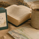 A green waxmelt gift box labeled 'The Last Chapter at Sea' sits beside an open book and a ceramic mug of coffee on a warm neutral surface, with dried grass and a shell and beach painting as styled props.