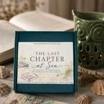 A square green gift box with a front label reading 'The Last Chapter at Sea' is displayed on a rustic tabletop with seashells, an open book, and a green owl-shaped candle holder in the background.