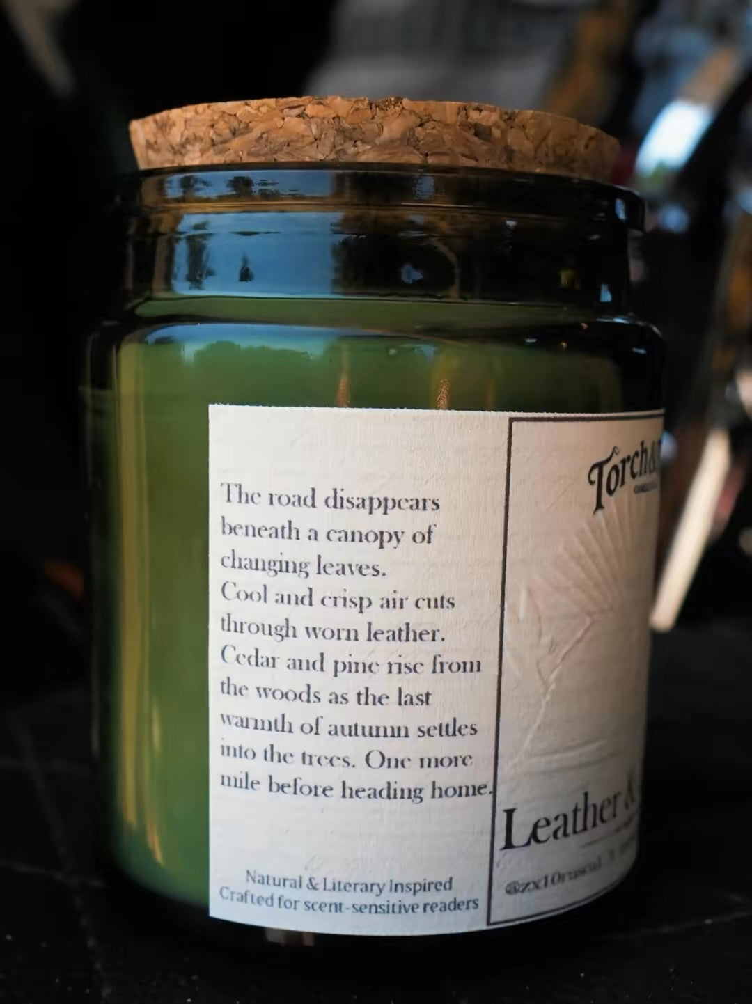 A jar candle with a cork stopper and a dark green glass body, featuring a white label with a quote about autumn and leather scents. The brand name 'Torch & Tome' is visible on the label.