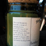 A jar candle with a cork stopper and a dark green glass body, featuring a white label with a quote about autumn and leather scents. The brand name 'Torch & Tome' is visible on the label.