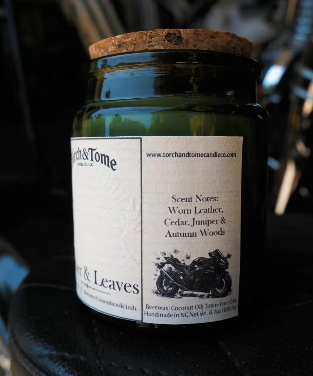 A dark green glass jar candle with a cork lid and a white paper label reading Torch & Tome, photographed close up against a dark, blurred background.