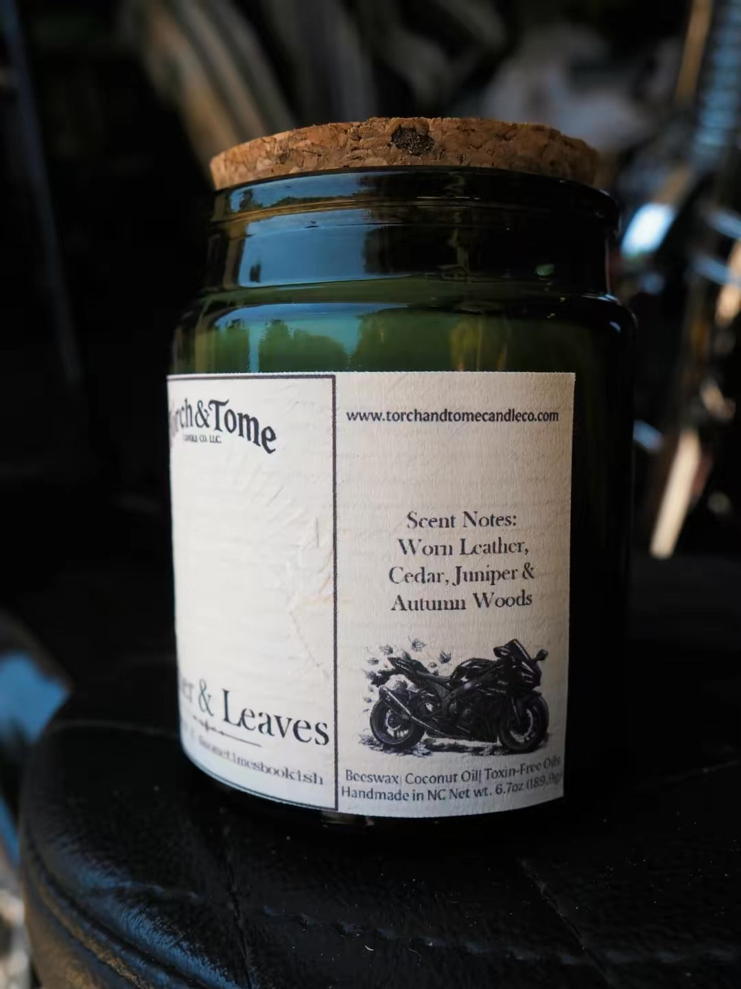 A dark green glass jar candle with a cork lid and a white paper label reading Torch & Tome, photographed close up against a dark, blurred background.