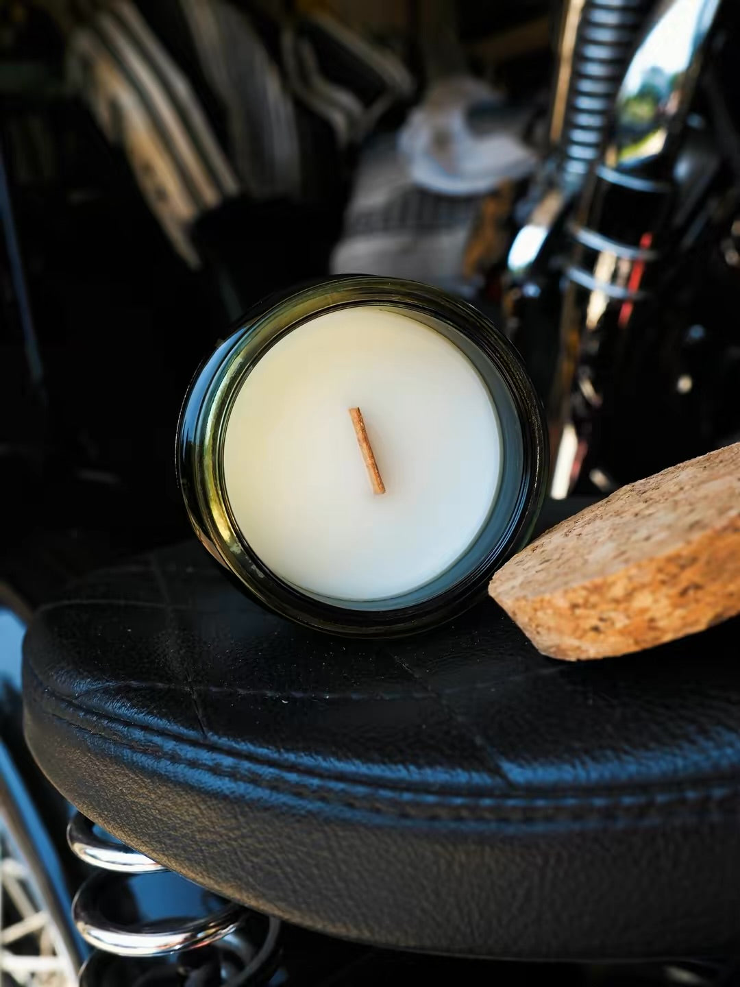 A jar candle with white wax and a single wick sits on a black leather seat next to a cork slice, with a blurred background showing a motorcycle or vehicle interior.