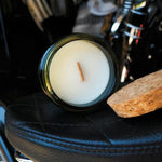A jar candle with white wax and a single wick sits on a black leather seat next to a cork slice, with a blurred background showing a motorcycle or vehicle interior.