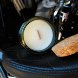 A jar candle with white wax and a single wick sits on a black leather seat next to a cork slice, with a blurred background showing a motorcycle or vehicle interior.
