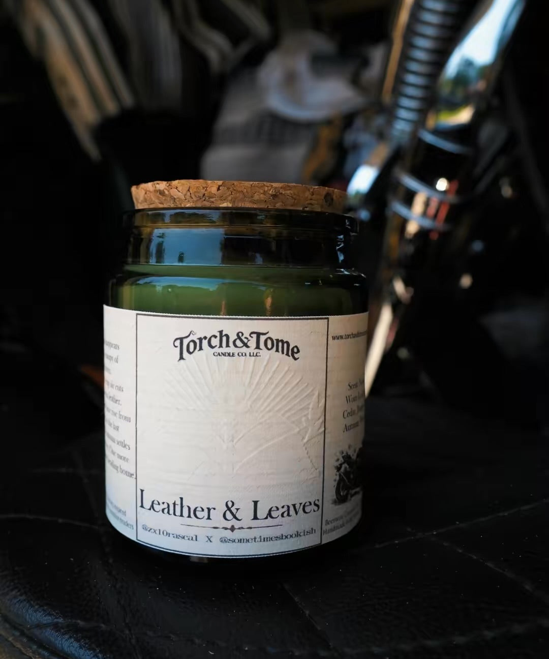A green glass jar candle with a cork lid and a white paper label reading 'Torch & Tome' and 'Leather & Leaves,' placed on a black leather surface with a blurred dark background.