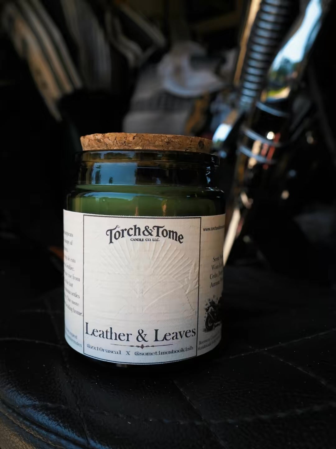 A green glass jar candle with a cork lid and a white paper label reading 'Torch & Tome' and 'Leather & Leaves,' placed on a black leather surface with a blurred dark background.