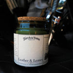 A green glass jar candle with a cork lid and a white paper label reading 'Torch & Tome' and 'Leather & Leaves,' placed on a black leather surface with a blurred dark background.