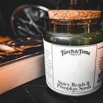 A candle in an amber glass jar with a cork stopper and a white label reading 'Torch & Tome' and 'Spicy Reads & Pumpkin Seeds,' styled on a dark surface beside an open book.