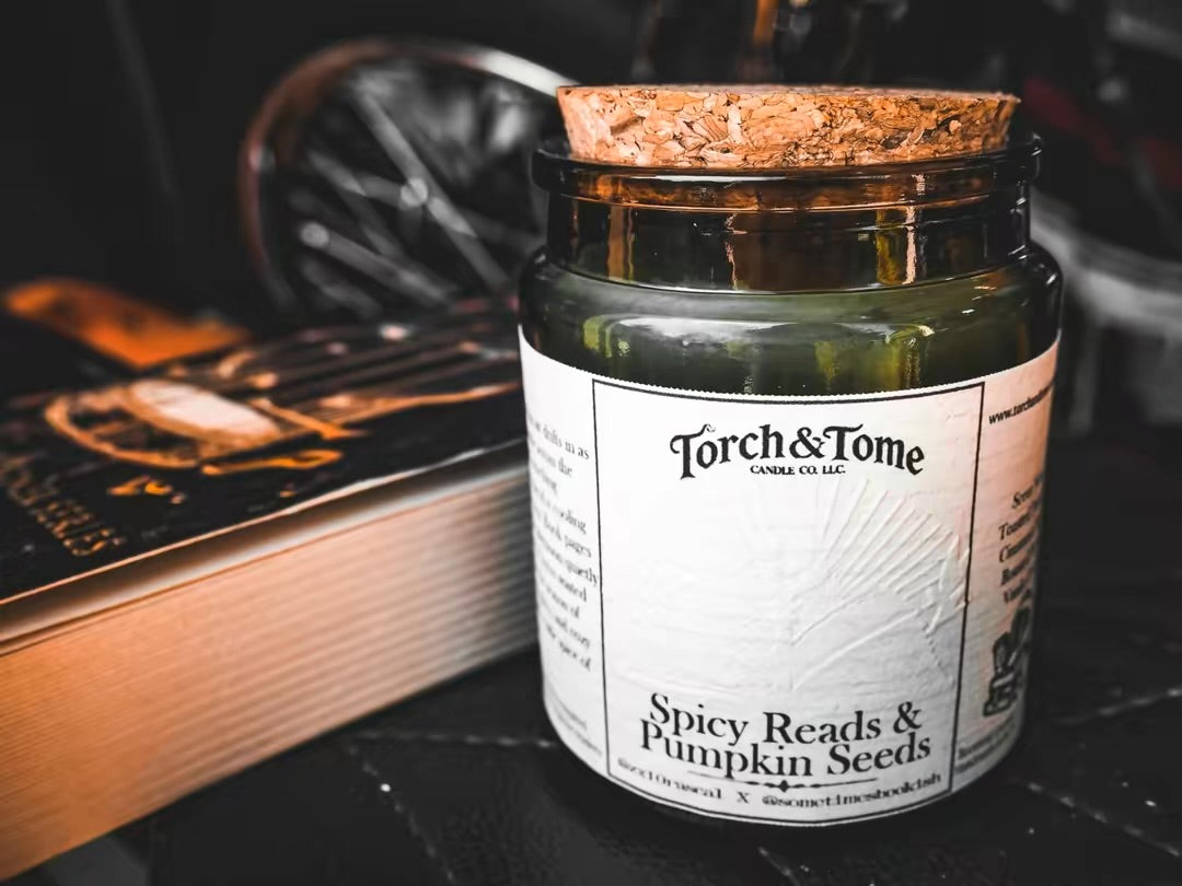A candle in an amber glass jar with a cork stopper and a white label reading 'Torch & Tome' and 'Spicy Reads & Pumpkin Seeds,' styled on a dark surface beside an open book.