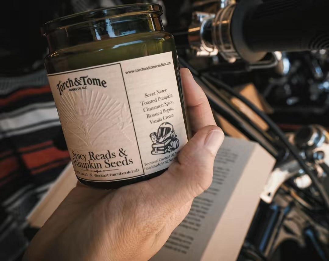 A hand holds a glass jar candle labeled 'Parch & Tome Spicy Reads & Pumpkin Seeds' in front of a motorcycle engine and open book, suggesting a cozy, autumnal theme.