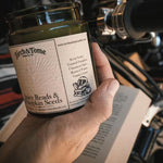 A hand holds a glass jar candle labeled 'Parch & Tome Spicy Reads & Pumpkin Seeds' in front of a motorcycle engine and open book, suggesting a cozy, autumnal theme.