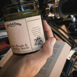 A hand holds a glass jar candle labeled 'Parch & Tome Spicy Reads & Pumpkin Seeds' in front of a motorcycle engine and open book, suggesting a cozy, autumnal theme.